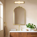 A bathroom features the LuxenHome Rêverie vintage gold accent metal frame arch mirror above a wooden vanity with a white countertop, adorned with a round green plant. Gold fixtures, such as a faucet and light fixture, harmonize with the beige walls.