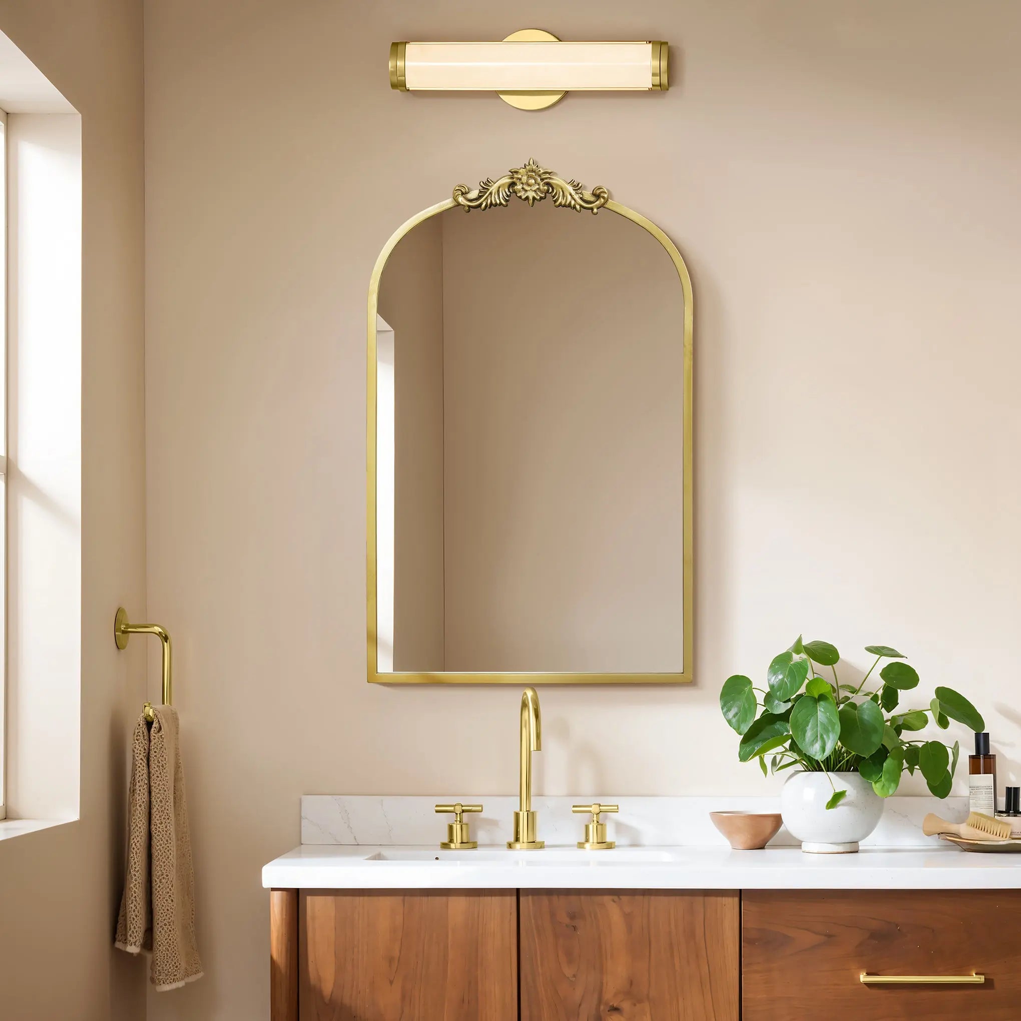 A bathroom features the LuxenHome Rêverie vintage gold accent metal frame arch mirror above a wooden vanity with a white countertop, adorned with a round green plant. Gold fixtures, such as a faucet and light fixture, harmonize with the beige walls.