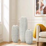 Three Modern geometric pattern resin tall vases by LuxenHome stand on a light wooden floor in a bright room. A beige sofa with a yellow cushion is partly visible on the right, with a framed abstract photo hanging above, adding artistic flair to the space.