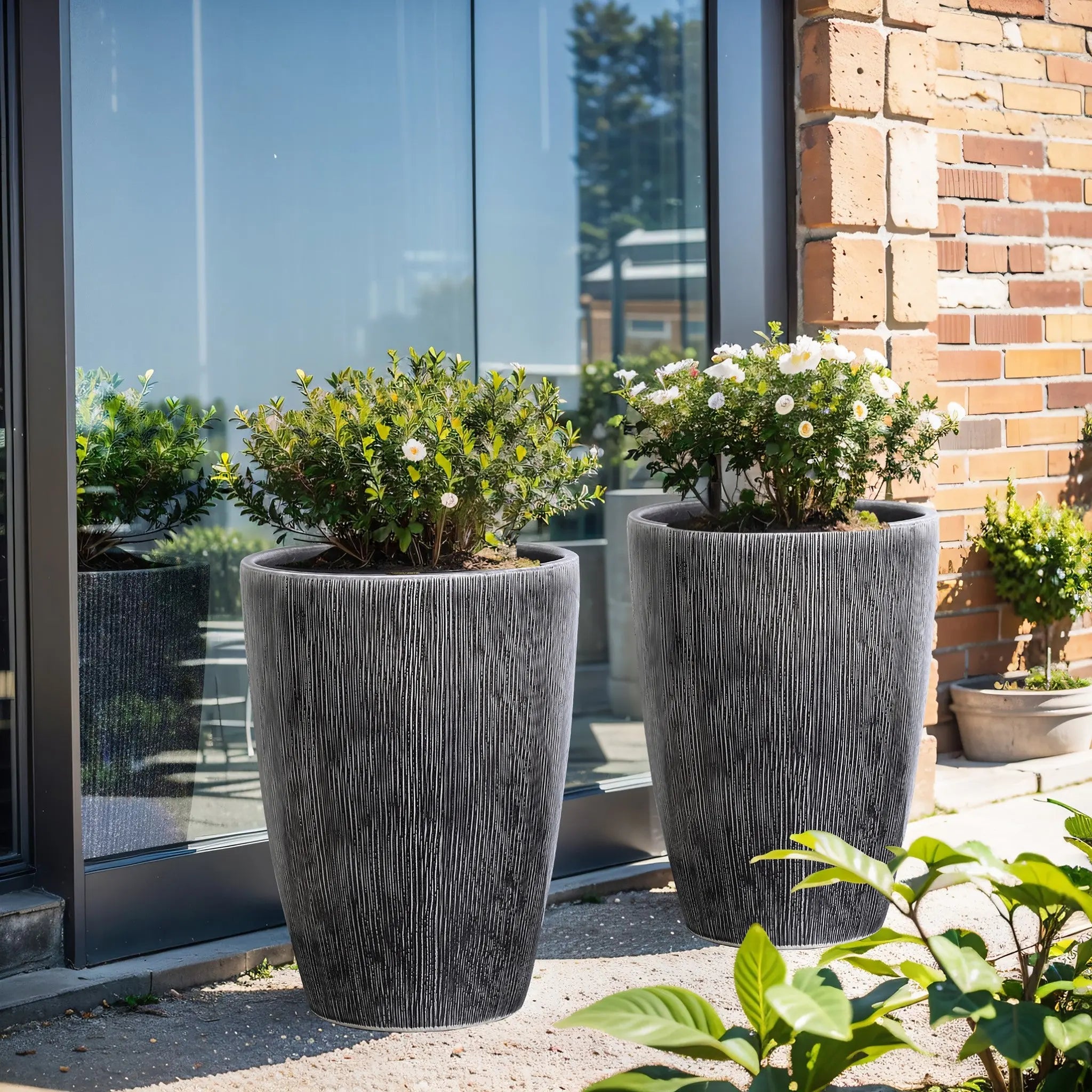 On the patio, two LuxenHomes Sea gray ripples glazed tall ceramic planters, 17 high, with lush green foliage and white flowers stand in front of a glass door and brick wall. These sleek black-and-white vertical striped planters have a few smaller plants visible in the background.