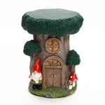 A decorative gnome tree house figurine featuring a tree stump house with a round window, door, two mushrooms, and a gnome beside it¡ªperfect for adding charm to your outdoor space decor. Set against a white background.