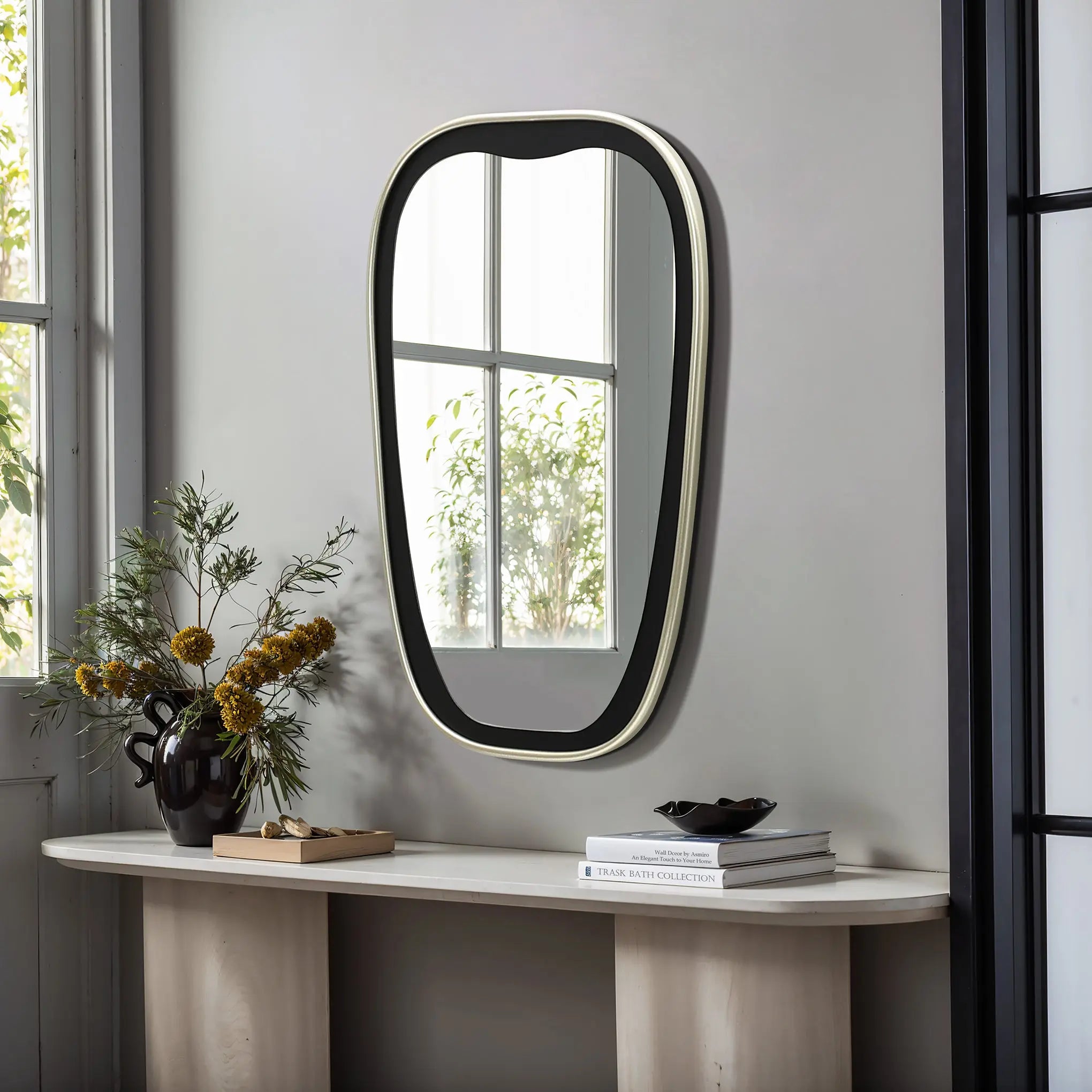 A contemporary mirror with an oblong shape and black frame is mounted above a light-colored console table. The table holds a black vase with flowers, stacked books, and a bowl. A window, offering glimpses of lush greenery, enhances the serene setup.