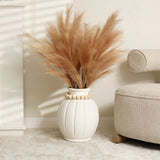 White vase with a modern round shape and beige pampas grass sits next to a textured cream sofa on a light carpeted floor, embodying minimalist design.