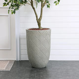 A sleek, modern minimalist design features a tall woven-patterned planter with a small tree, set against a white paneled wall on the dark tiled floor.