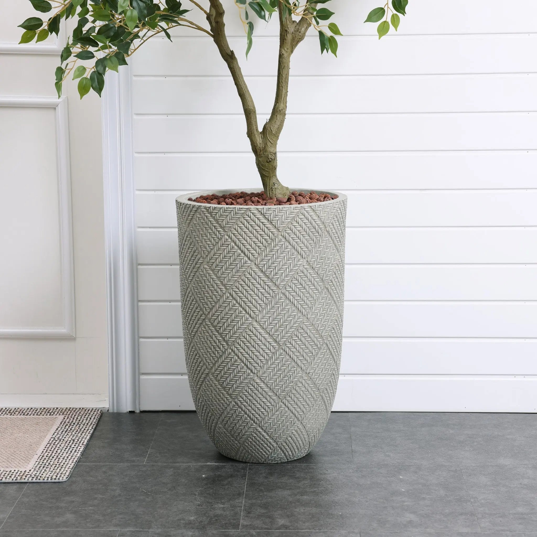 A sleek, modern minimalist design features a tall woven-patterned planter with a small tree, set against a white paneled wall on the dark tiled floor.