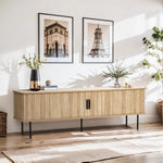 A modern living room features a light wood sideboard, highlighted by two framed architectural prints above, a sleek black vase, a stack of books, and a potted plant. An oak TV stand with tambour doors seamlessly blends in while natural light brightens the space.