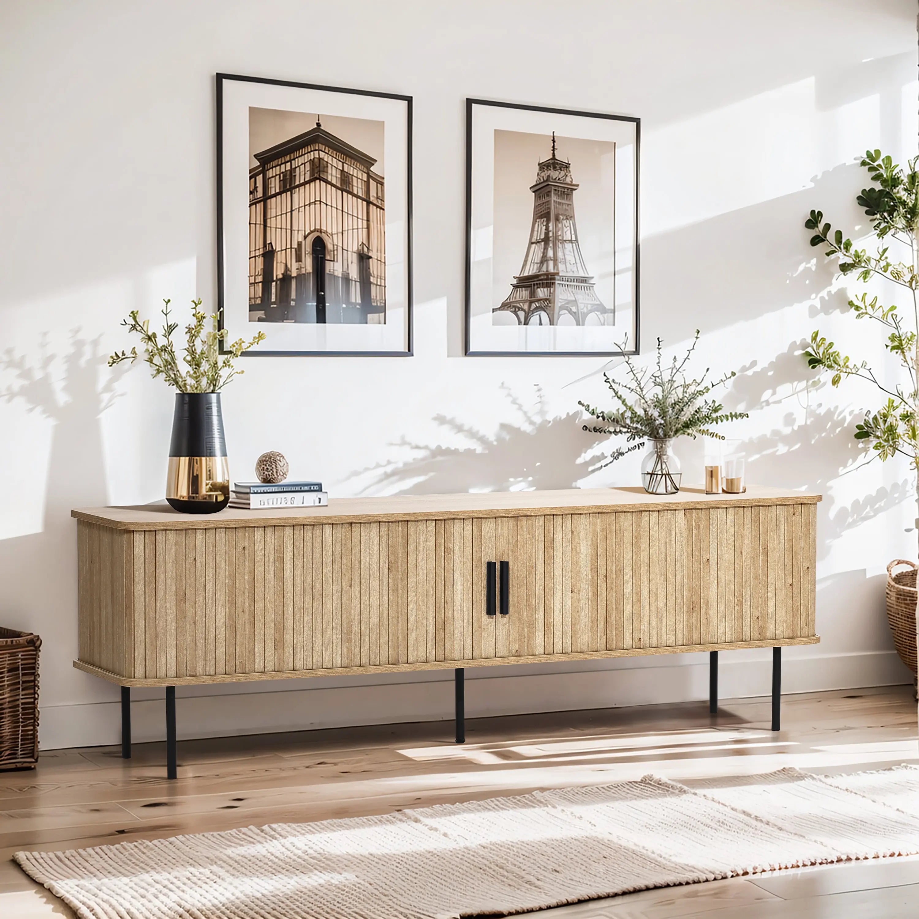A modern living room features a light wood sideboard, highlighted by two framed architectural prints above, a sleek black vase, a stack of books, and a potted plant. An oak TV stand with tambour doors seamlessly blends in while natural light brightens the space.