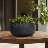 A LuxenHome Black beehive pattern round tabletop planter, 17.5 wide, holds lush green ivy on a wooden patio table, surrounded by partial views of pillars and greenery in the background.