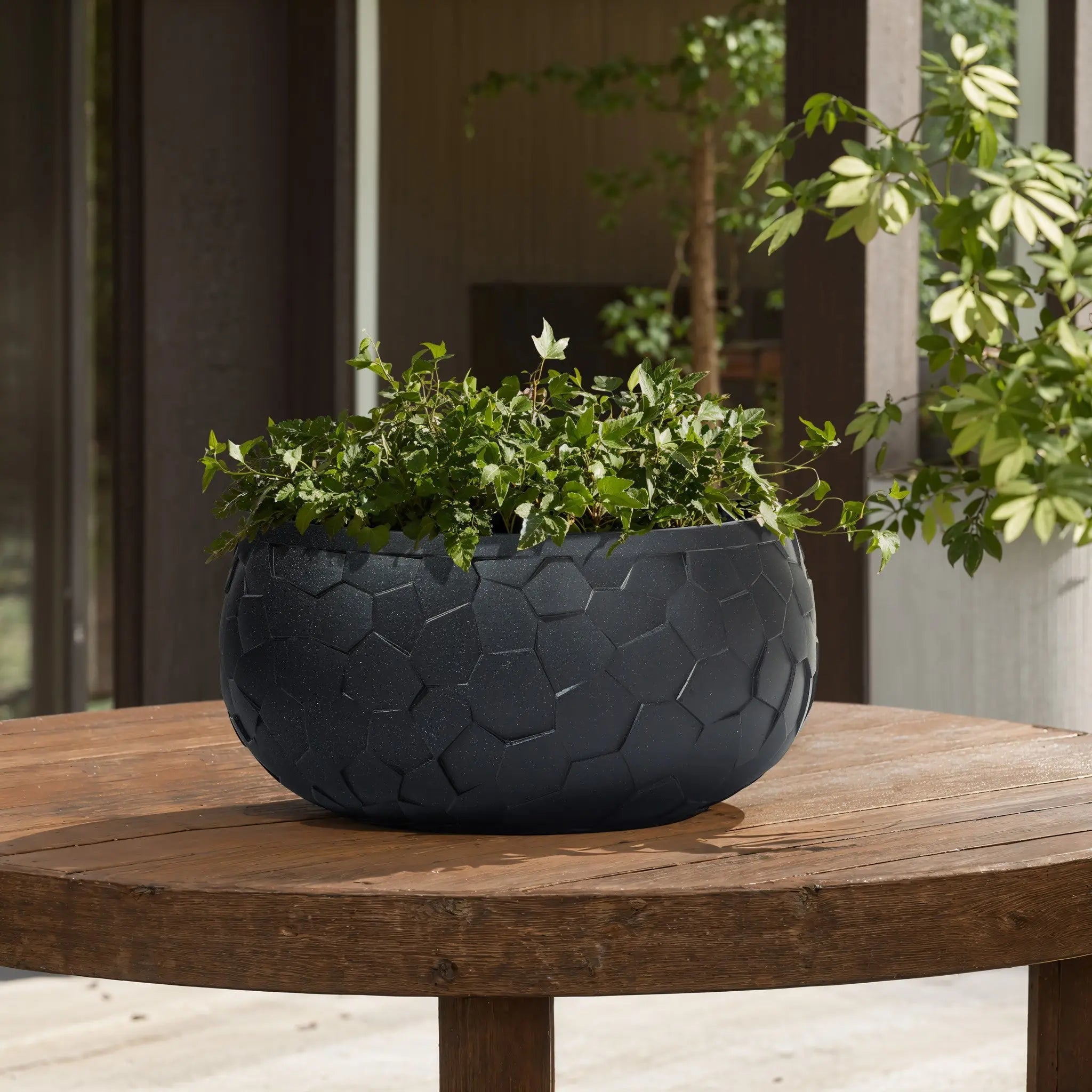 A LuxenHome Black beehive pattern round tabletop planter, 17.5 wide, holds lush green ivy on a wooden patio table, surrounded by partial views of pillars and greenery in the background.