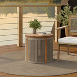 A wooden patio set crafted from natural acacia wood features a round table and chair on a hand-woven rope rug, overlooking a serene lake. The drum-shaped end table holds a small plant and a cup, inviting relaxation in natures embrace.