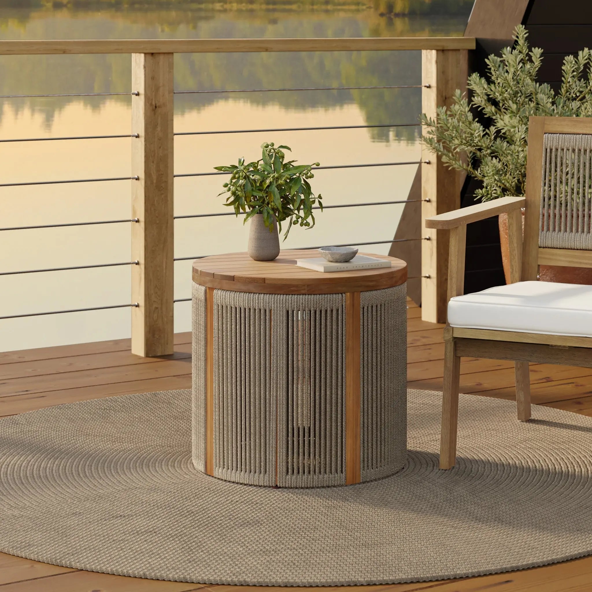 A wooden patio set crafted from natural acacia wood features a round table and chair on a hand-woven rope rug, overlooking a serene lake. The drum-shaped end table holds a small plant and a cup, inviting relaxation in natures embrace.