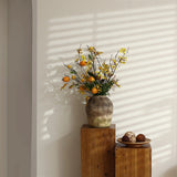 A rustic terracotta vase with yellow flowers and orange fruit sits on a wooden pedestal next to a bowl of nuts. Its handcrafted weathered finish complements the sunlight, which creates striped shadows on the wall.