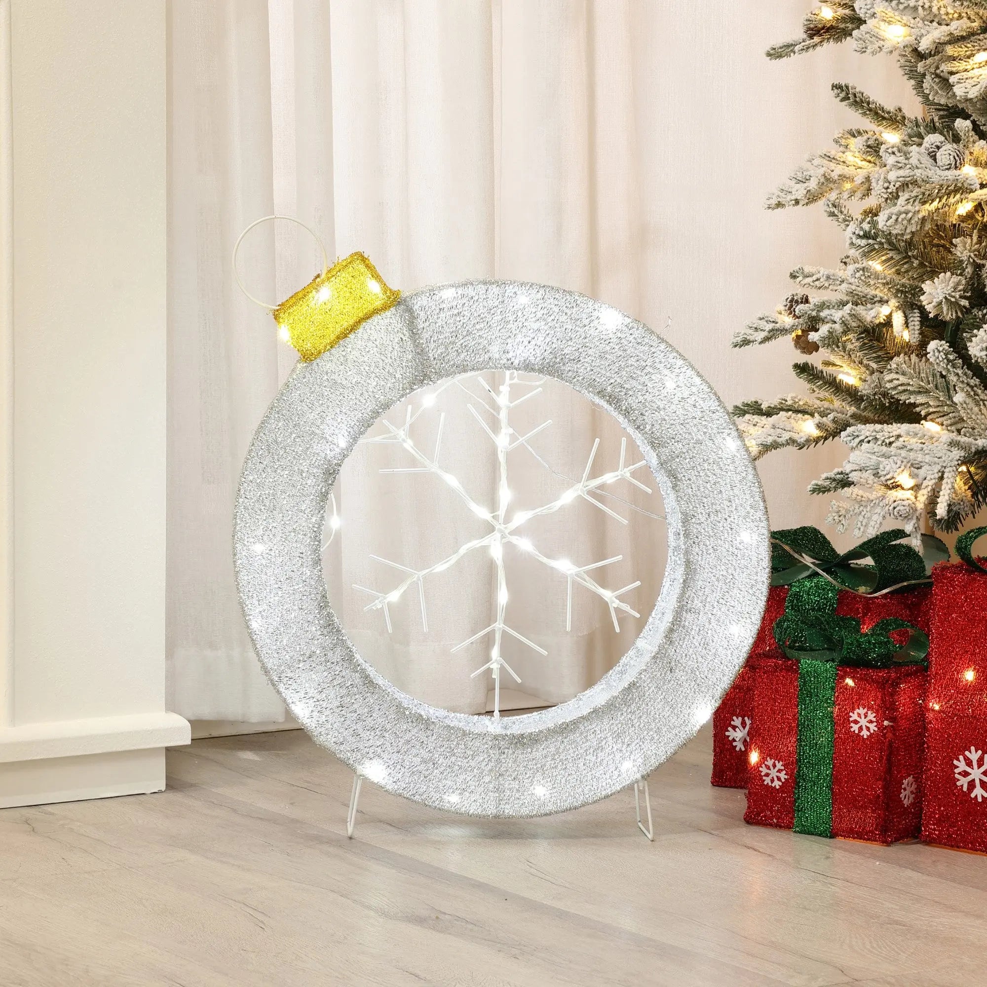 A large silver ornament with white lights and a snowflake design complements a lit Christmas tree and red gift boxes, perfect for adding festive charm to your holiday decoration.