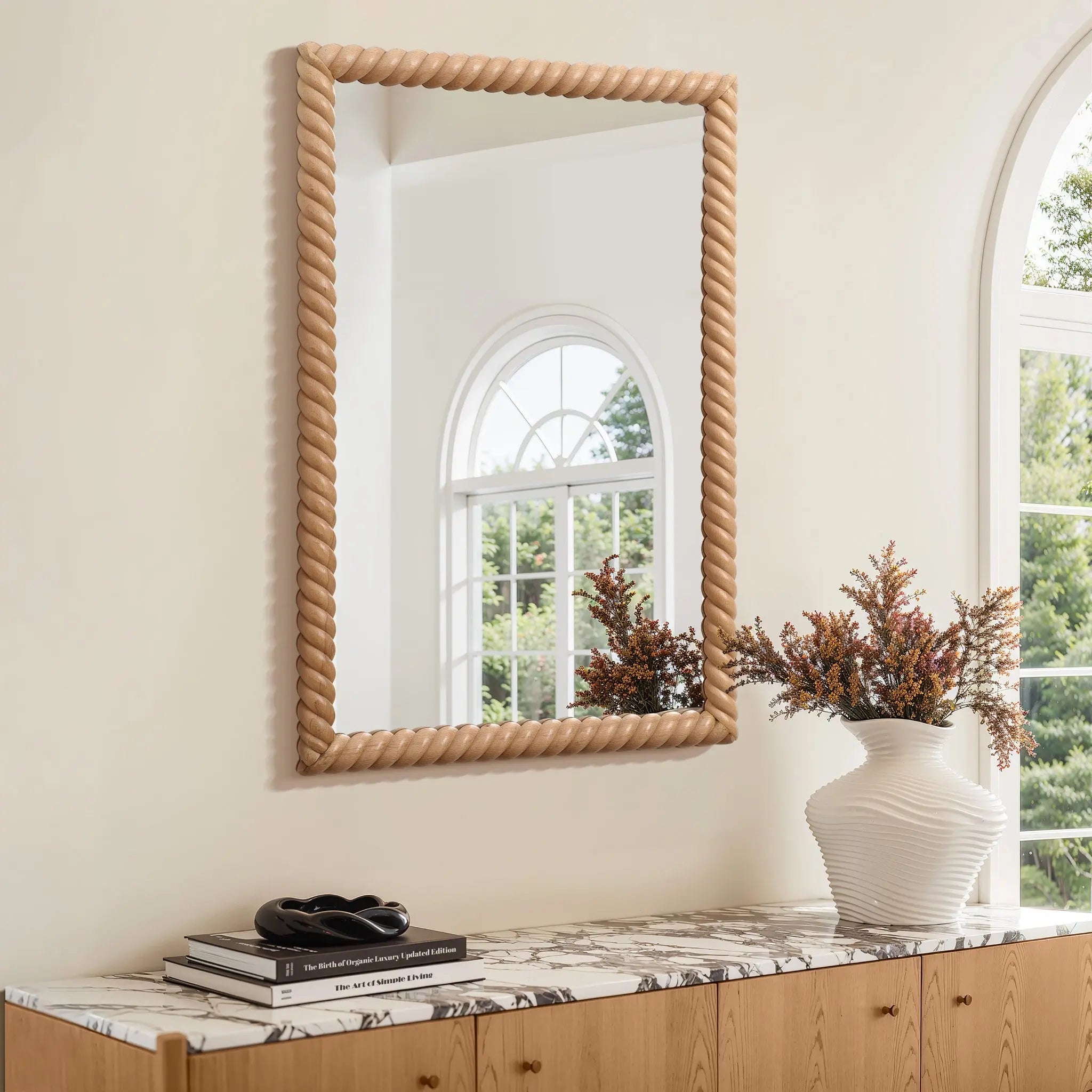An elegant decorative wall mirror with a twisted frame hangs above a wooden console table. The table, adorned with an opulent marble top, showcases a stack of books and a vase with dried flowers, creating an impeccable touch of elegant home decor.