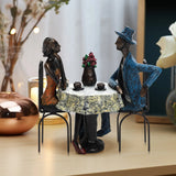 Two decorative sculptures of a modern brown resin couple sitting at a table with coffee and a vase of flowers, placed on a wooden surface with a blurred background. This charming piece captures romantic moments, making it an ideal wedding gift.