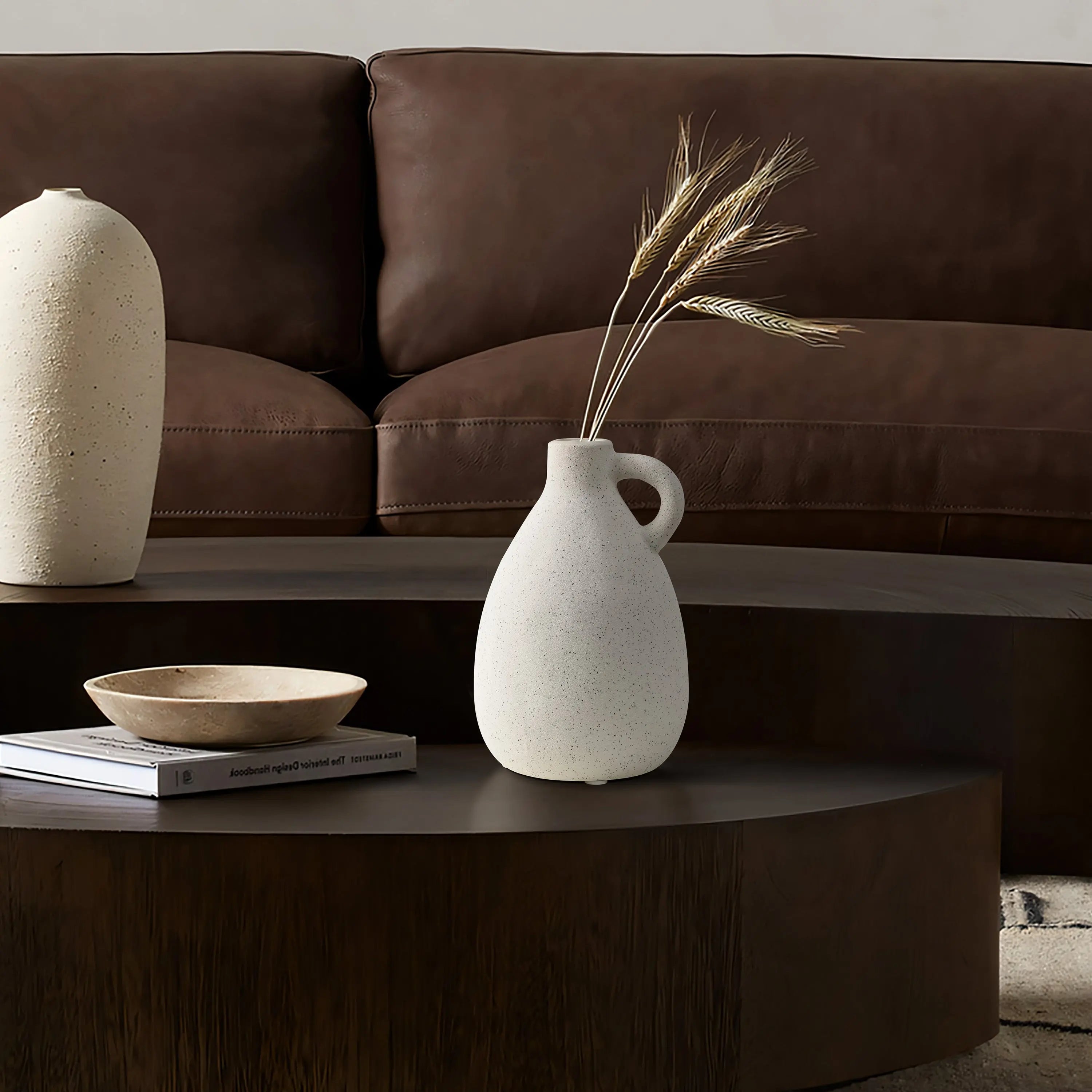 A minimalist living room scene featuring a brown leather sofa, a vintage vase with dried stems beside terracotta accents, a wooden coffee table, and stacked books perfectly captures the essence of thoughtful home decor.