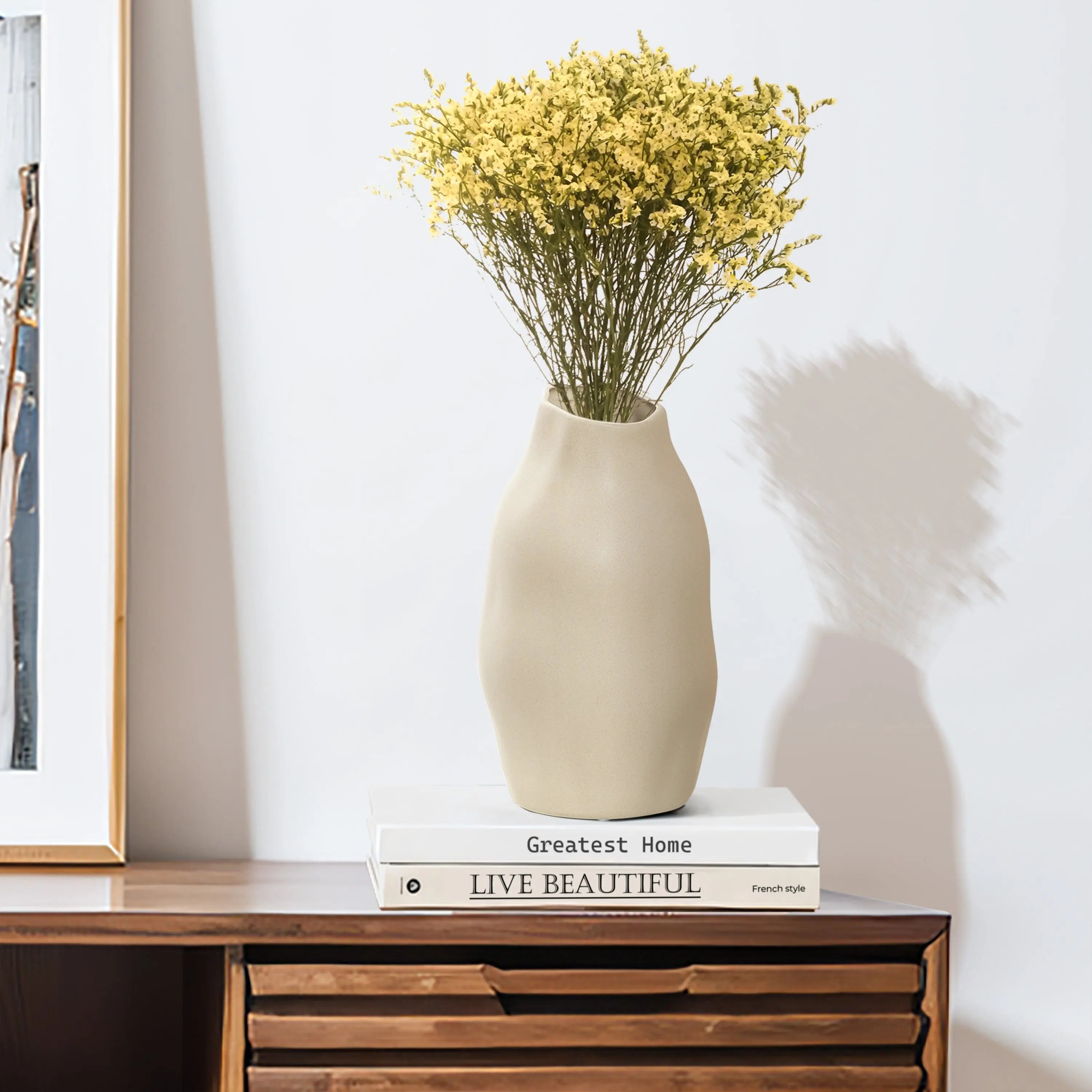 An ivory white ceramic vase with yellow flowers sits as an accent piece atop a stack of books titled Greatest Home and Live Beautiful on a wooden table.