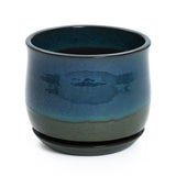 A round, glossy blue-green ceramic planter with drainage holes, it showcases a dark base and comes with a circular saucer at the bottom.