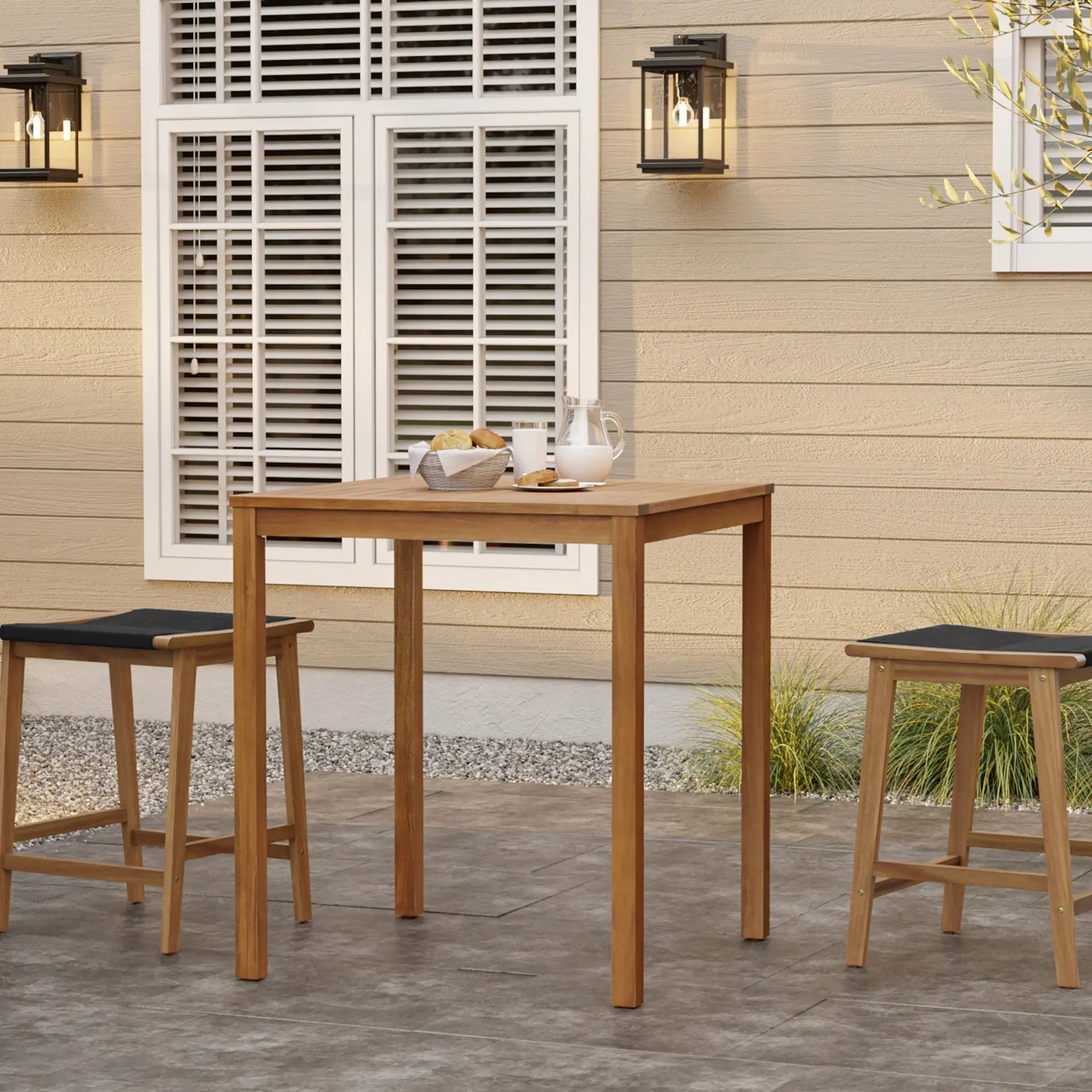 Two wooden stools and a charming square table crafted from FSC-Certified Acacia Wood are on a patio in front of a beige building with a window. Lanterns are mounted on the wall, and plants flourish in the background, creating an inviting outdoor space.