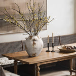 A large handcrafted vintage vase with branches and yellow flowers sits on a wooden dining table, accompanied by three lit candles in black holders and a wicker bowl with decorative objects. This versatile home decor piece adds charm to any setting.