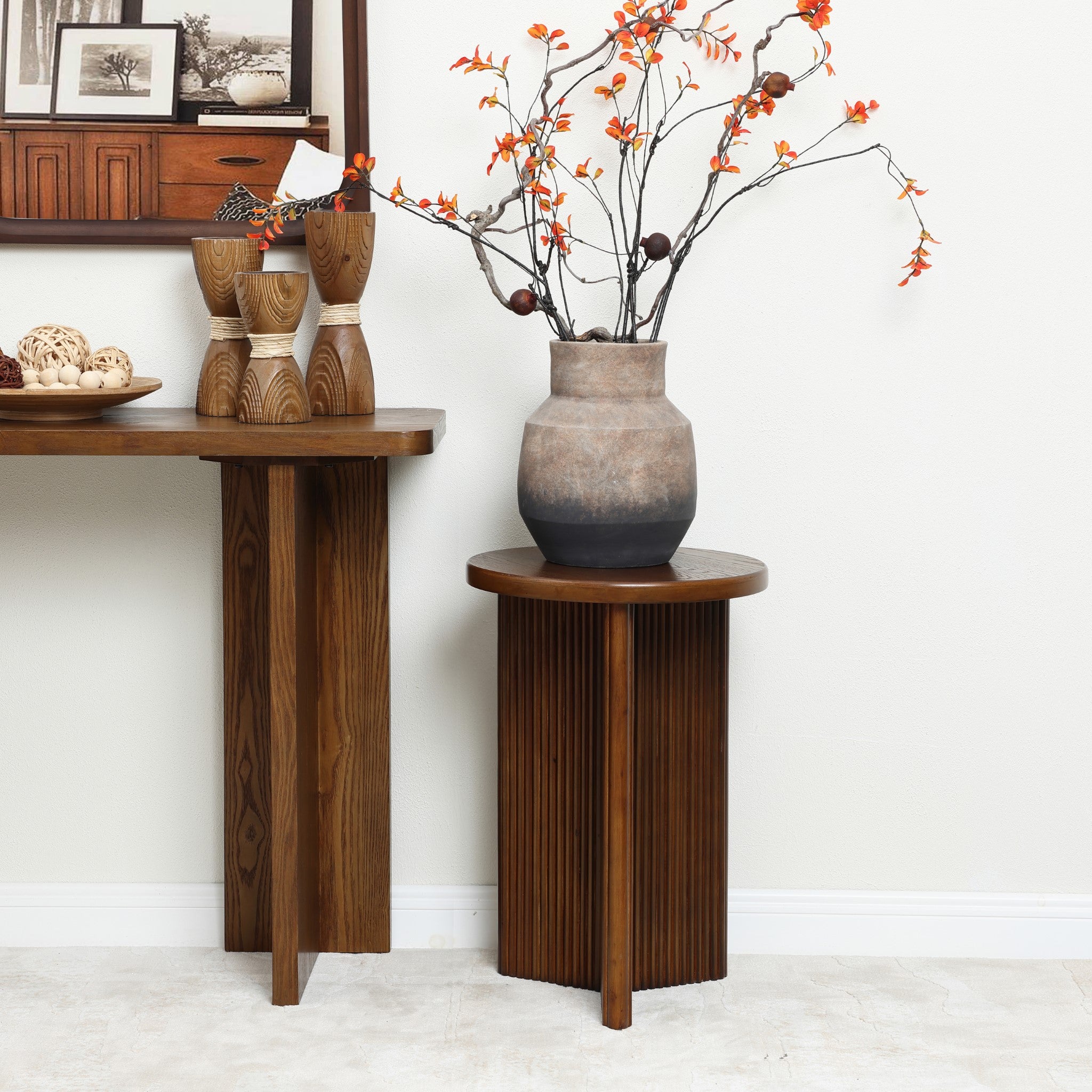 A wooden table with decorative wooden figurines and a ceramic vase filled with orange flowers sits prominently. Nearby, a mid-century modern side table features a similar vase with branches. Framed pictures adorn the wall shelf, completing the charming scene.