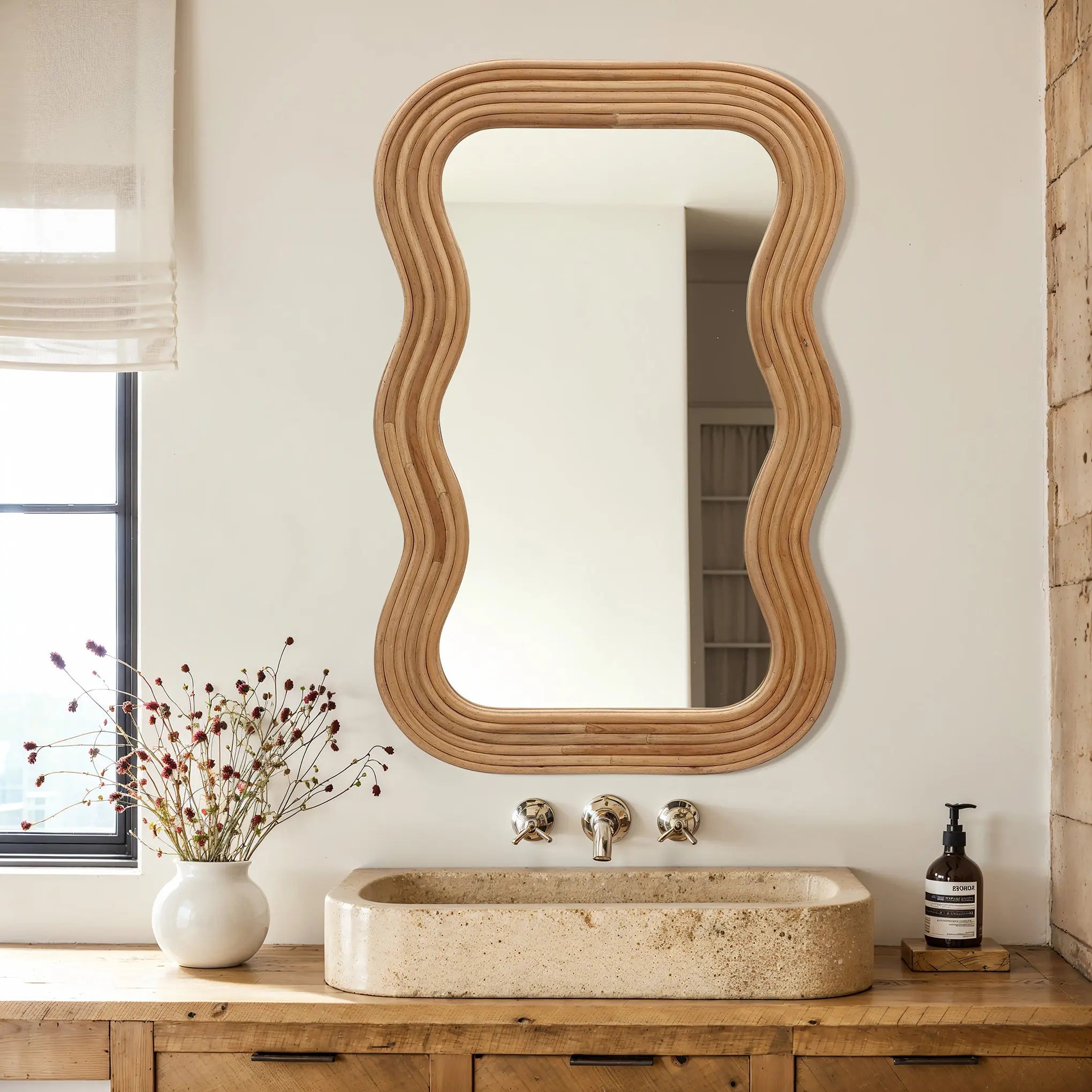 A cork wood-framed, wavy mirror hangs above a beige stone sink on a wooden counter, complemented by a vase of dried flowers and a soap dispenser, creating a modern vibe.