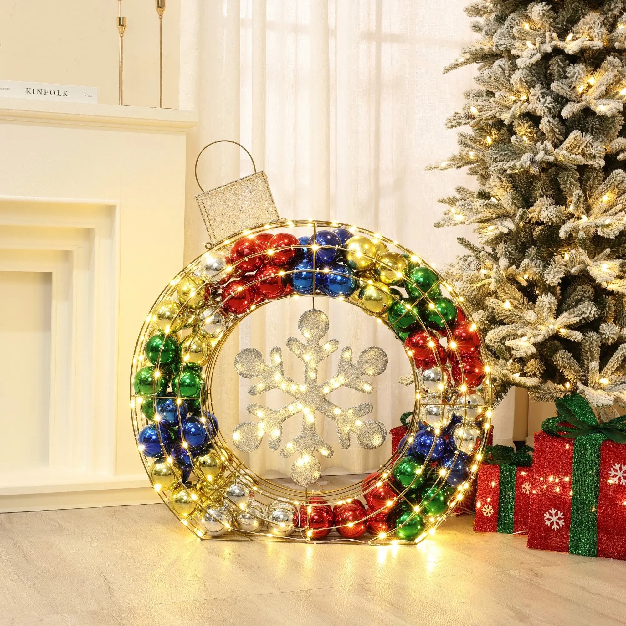 A large ornament-shaped holiday decoration with colorful lights and a snowflake design sits next to a snow-dusted Christmas tree and wrapped presents.