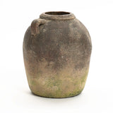 An aged, round clay jar with a small spout and weathered surface, showing signs of moss and wear, this rustic terracotta vase embodies a handcrafted, weathered finish suitable for both indoor and outdoor display.