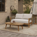 Outdoor patio with an Outdoor Acacia Wood Rope Sides Loveseat with white cushions, a matching coffee table set holding a teapot and cup, rug, umbrella, lush plants, and a stone wall backdrop.