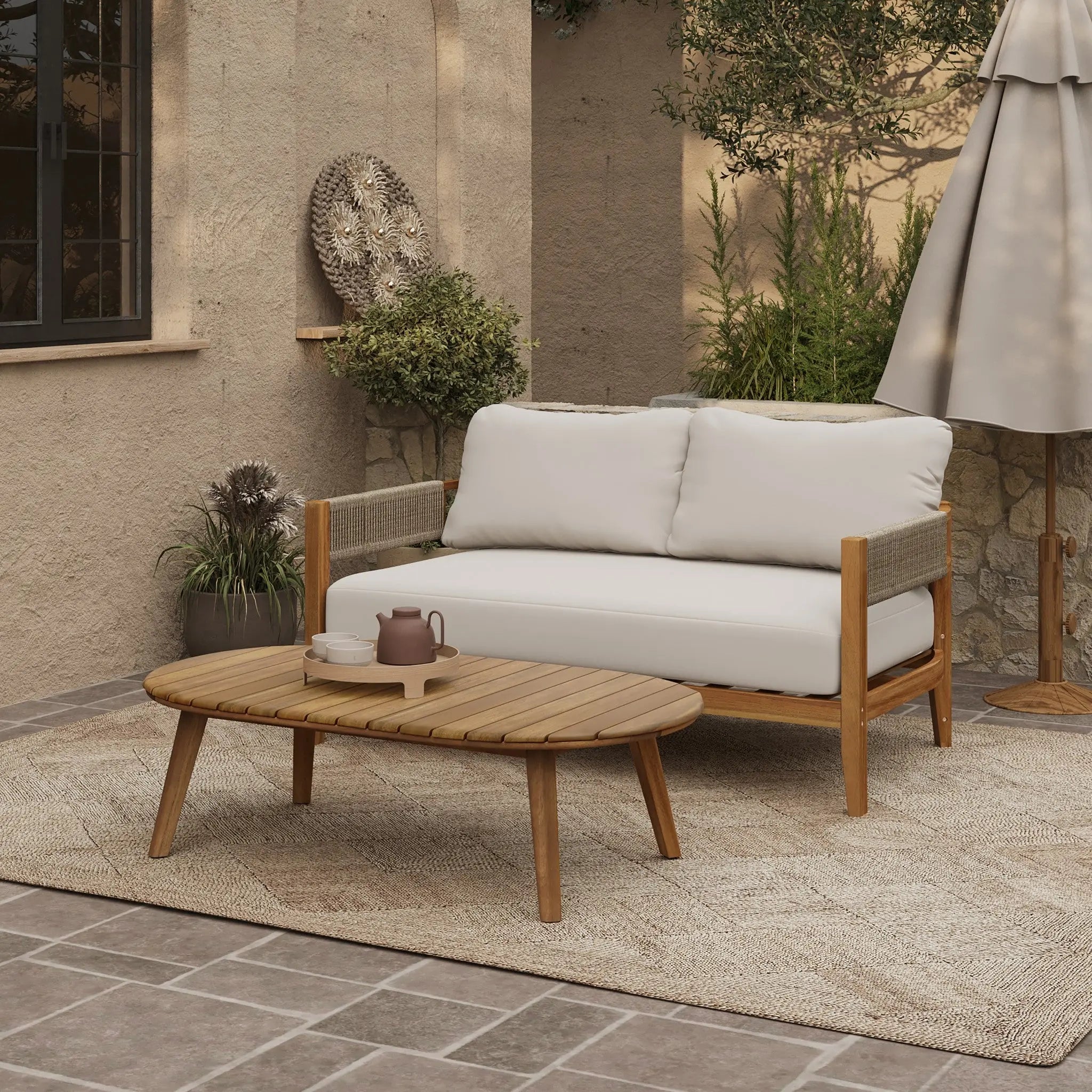 Outdoor patio with an Outdoor Acacia Wood Rope Sides Loveseat with white cushions, a matching coffee table set holding a teapot and cup, rug, umbrella, lush plants, and a stone wall backdrop.