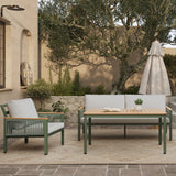 Outdoor patio featuring a wooden table and two cushioned chairs with plush cushions beneath a cream-colored umbrella. An outdoor loveseat sofa adds comfort, while plants and a large tree provide natural beauty near the stone wall with its light fixture on the left.