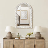 An arched rectangular mirror with a patterned frame elegantly hangs above a sideboard. Two lamps, a small plant, and glassware are neatly arranged below. Reflections of the staircase and wall art add to the charm of this home decoration.
