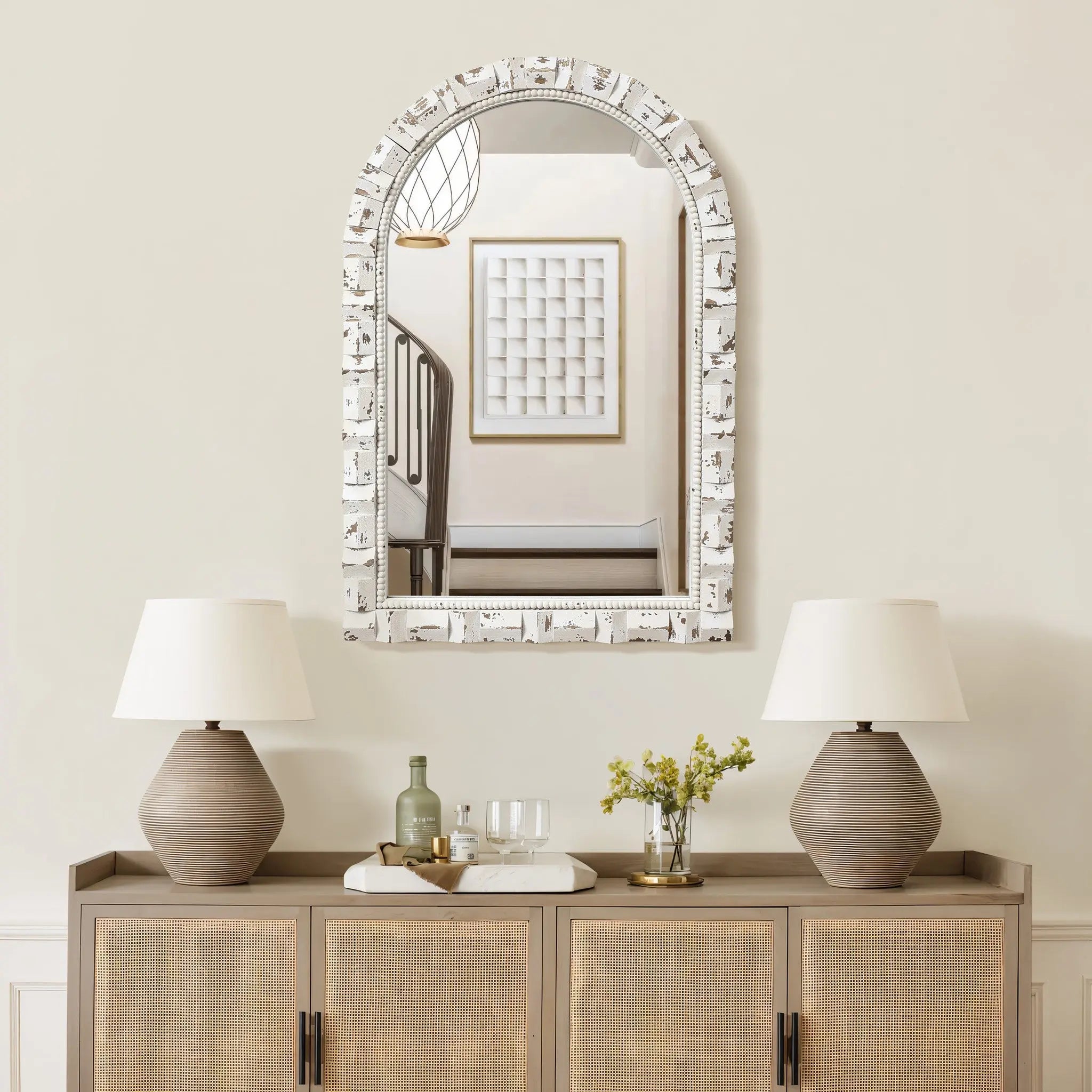 An arched rectangular mirror with a patterned frame elegantly hangs above a sideboard. Two lamps, a small plant, and glassware are neatly arranged below. Reflections of the staircase and wall art add to the charm of this home decoration.