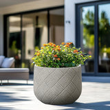 A durable MgO material planter with a woven pattern and blooming orange flowers is placed on a sunny patio near glass doors.