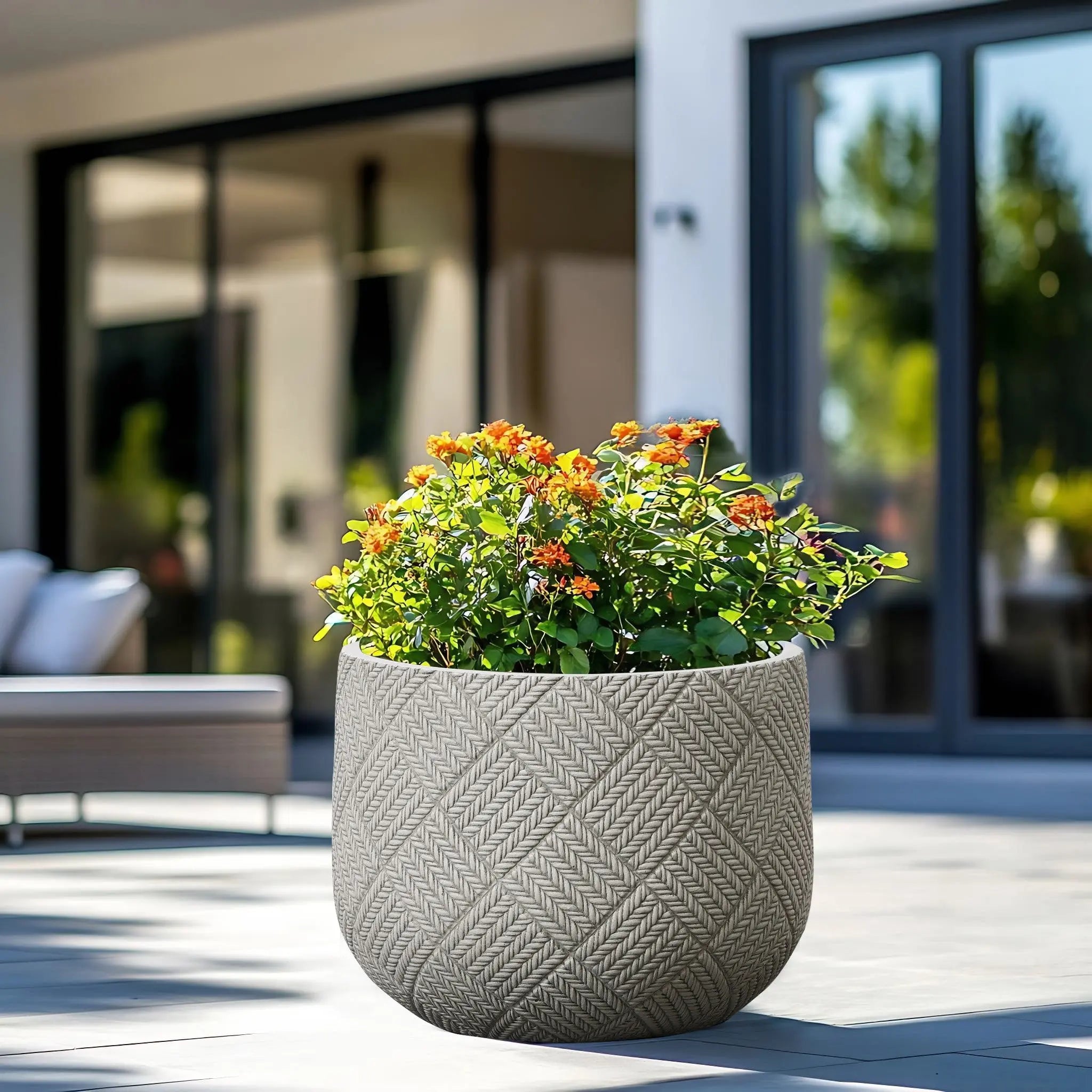 A durable MgO material planter with a woven pattern and blooming orange flowers is placed on a sunny patio near glass doors.