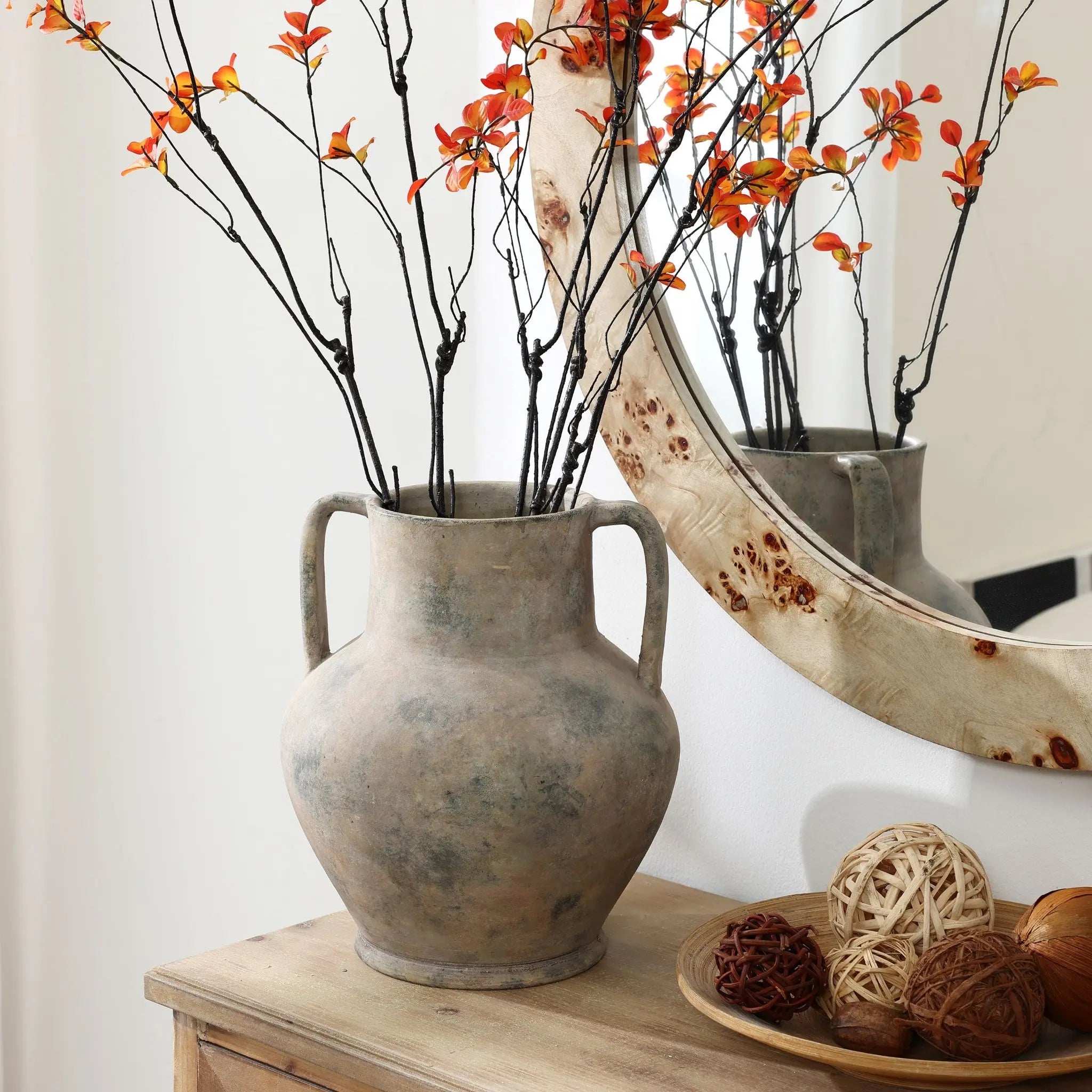 A LuxenHome 12.1 h mottled grey terracotta urn vase with handles holds branches of small orange flowers on a wooden table beside a large round mirror, accompanied by rustic decorative balls made of natural materials.
