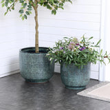 Two Marina gray glazed round ceramic planters from LuxenHome, featuring textured designs, sit on a dark tiled floor. The 13.98 h planter holds a small tree, while the smaller one is filled with colorful flowers and greenery; a textured rug is partially visible on the right.