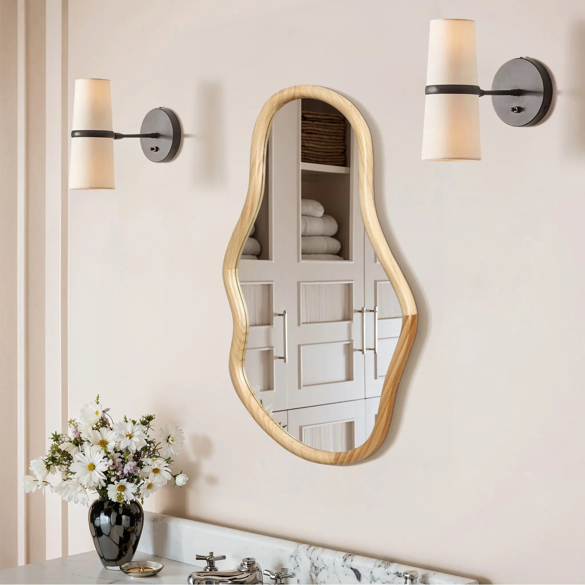 An asymmetrical mirror crafted from natural pine wood makes a decorative statement as it hangs on the wall with two sconces. Below, a marble countertop holds a vase of white flowers, while the mirror’s irregular oval shape reflects shelves stacked with towels.