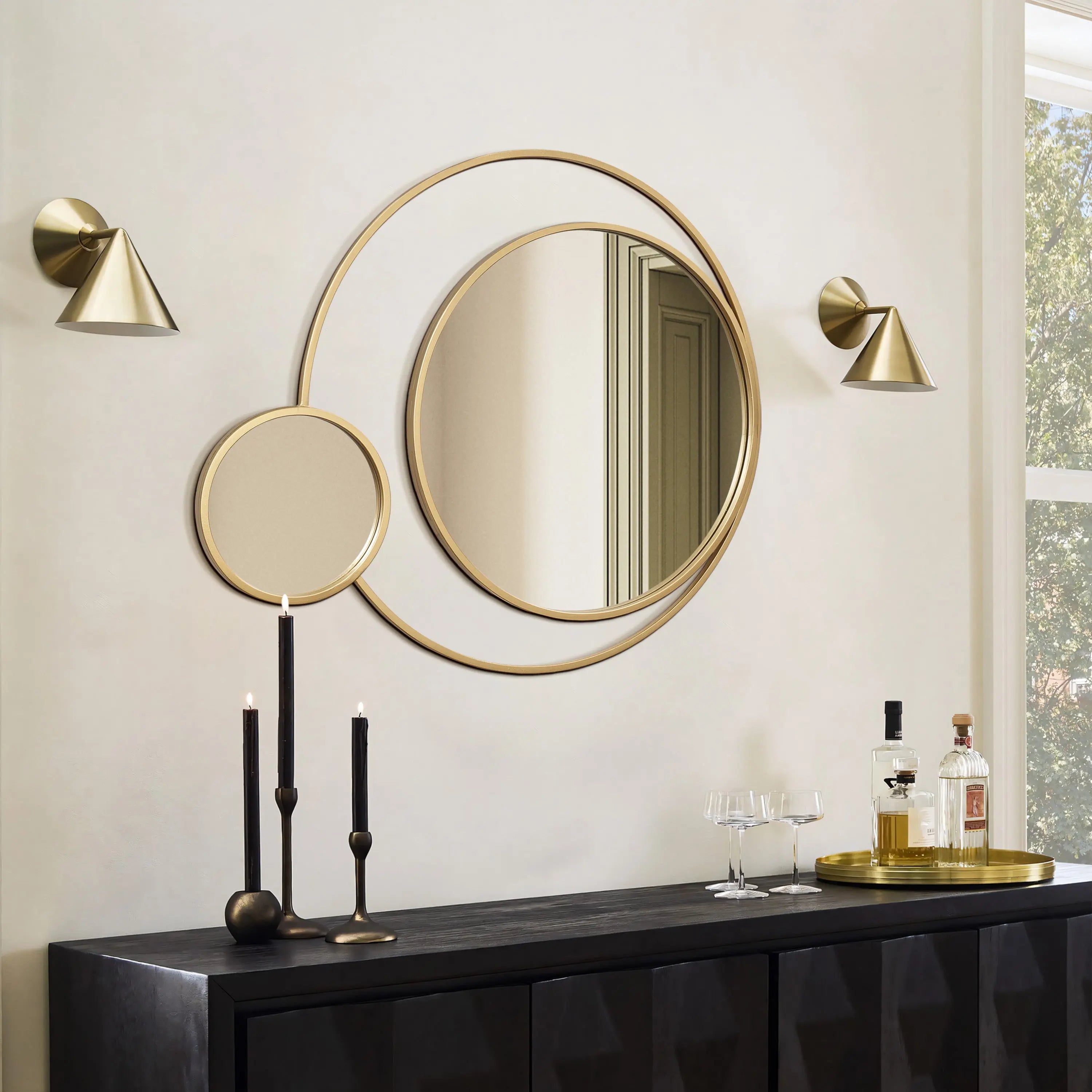 Wall-mounted decorative mirrors with golden frames are set against a light background, flanked by two small wall lights. A black sideboard below holds tall candles, glasses, and bottles on a tray, exuding modern elegance.