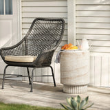 Wicker chair with a cushion beside a travertine-style side table holding a teapot and croissants on a plate, set against a white paneled wall.