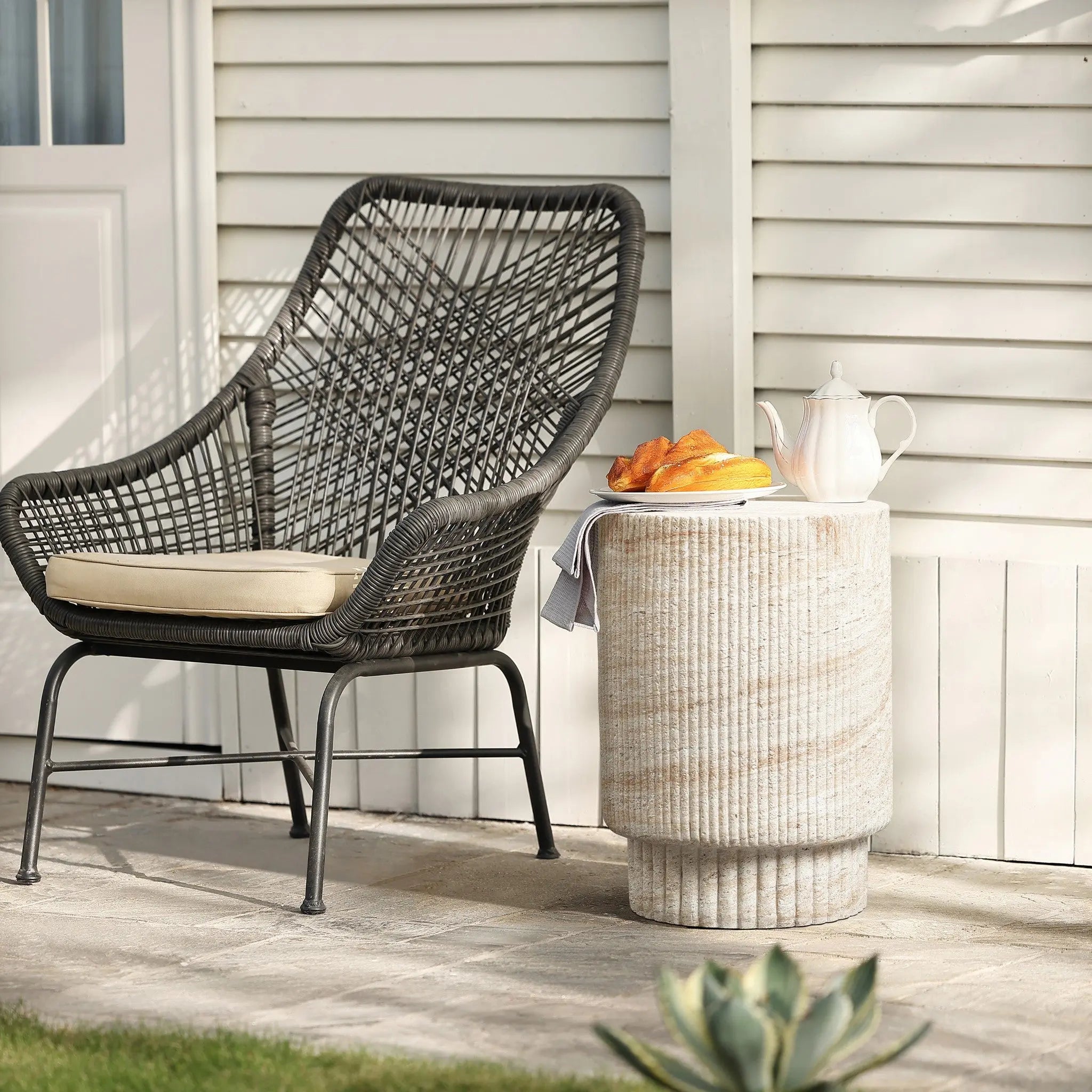 Wicker chair with a cushion beside a travertine-style side table holding a teapot and croissants on a plate, set against a white paneled wall.