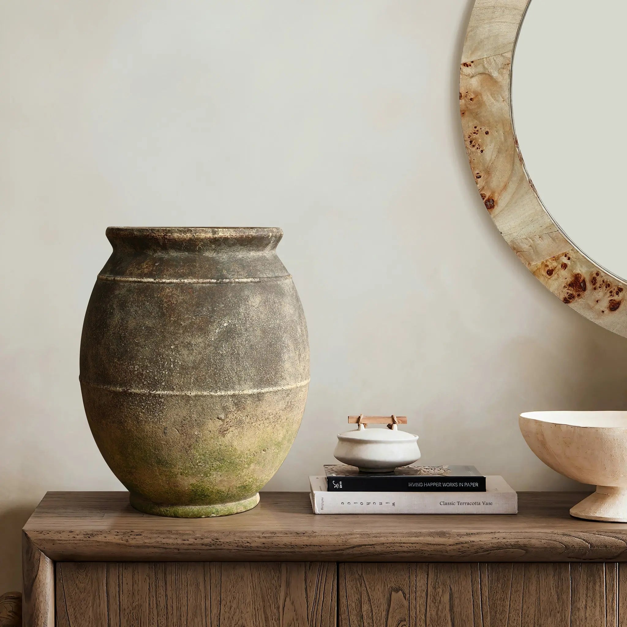A rustic ginger root vase on a wooden sideboard beside two books, a small dish, and a round mirror on the wall.