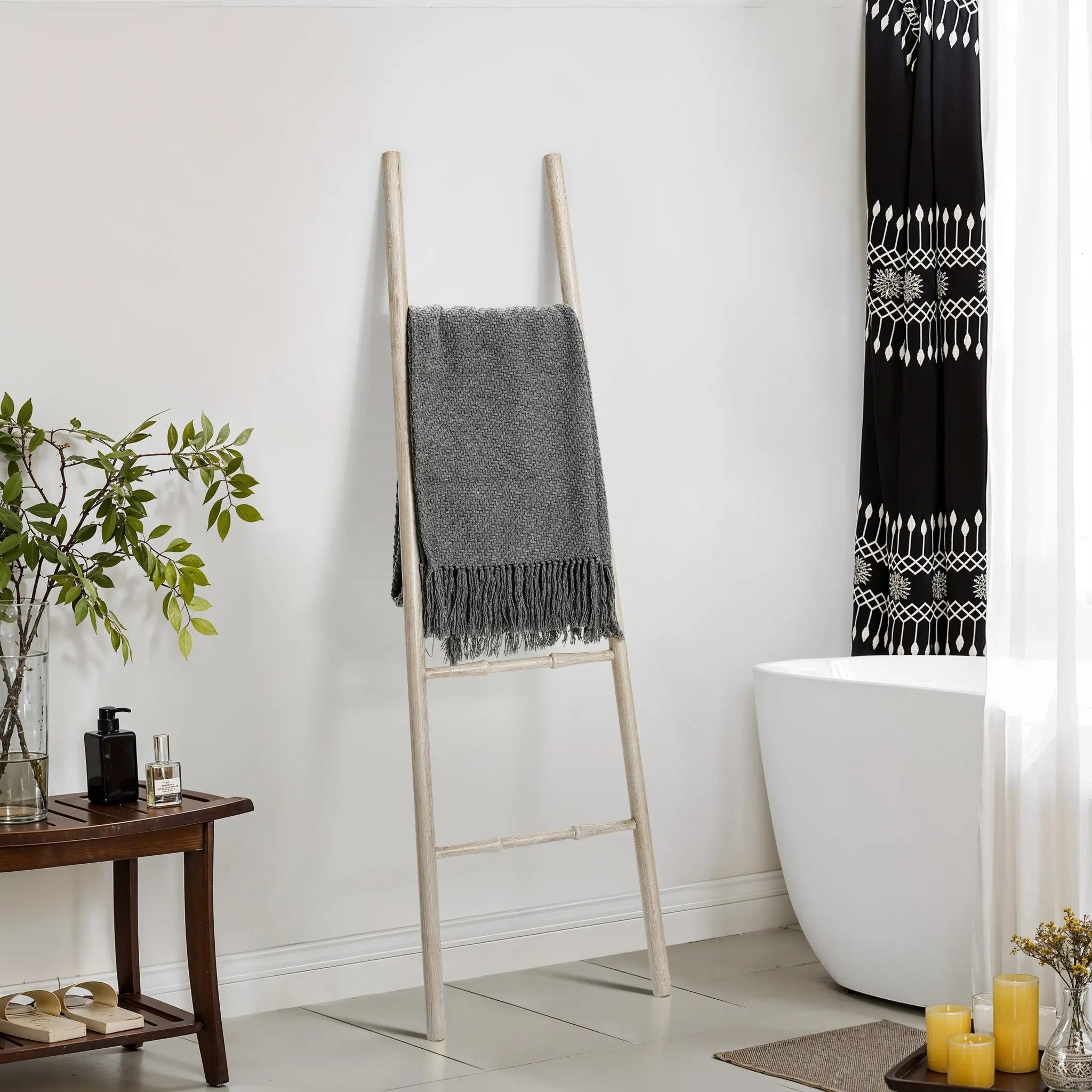 A rustic blanket ladder crafted from birch wood holds a gray fringed throw beside a white bathtub. Nearby, a wooden table adorned with plants, candles, and toiletries exudes charm, while a patterned black and white curtain adds contrast.