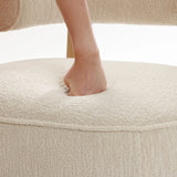 A persons foot presses down on a cream-hued Rio upholstered accent chair by LuxenHome, creating an indentation. The plush texture enhances a contemporary living space, with the plain white background highlighting the chairs elegant simplicity.