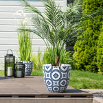 A modern planter made of MgO material, featuring a potted palm, sits gracefully in a decorative blue and white design on the wooden deck. Nearby, two black lanterns with candles add charm, while another similar planter with lush greenery enriches the background.