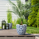 A modern planter made of MgO material, featuring a potted palm, sits gracefully in a decorative blue and white design on the wooden deck. Nearby, two black lanterns with candles add charm, while another similar planter with lush greenery enriches the background.