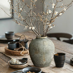 A LuxenHome Rustic gold and brown terracotta bud vase, 8.7 h, sits on a wooden table adorned with delicate white blossoms. It is surrounded by black ceramic cups and small bowls, creating an elegant dining setup that adds an antique distressed look to any home decor.