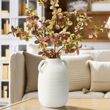 A textured ceramic vase with handles holds a leafy branch with brown and green leaves. It exudes rustic charm as it sits on a wooden table in the cozy living room setting.