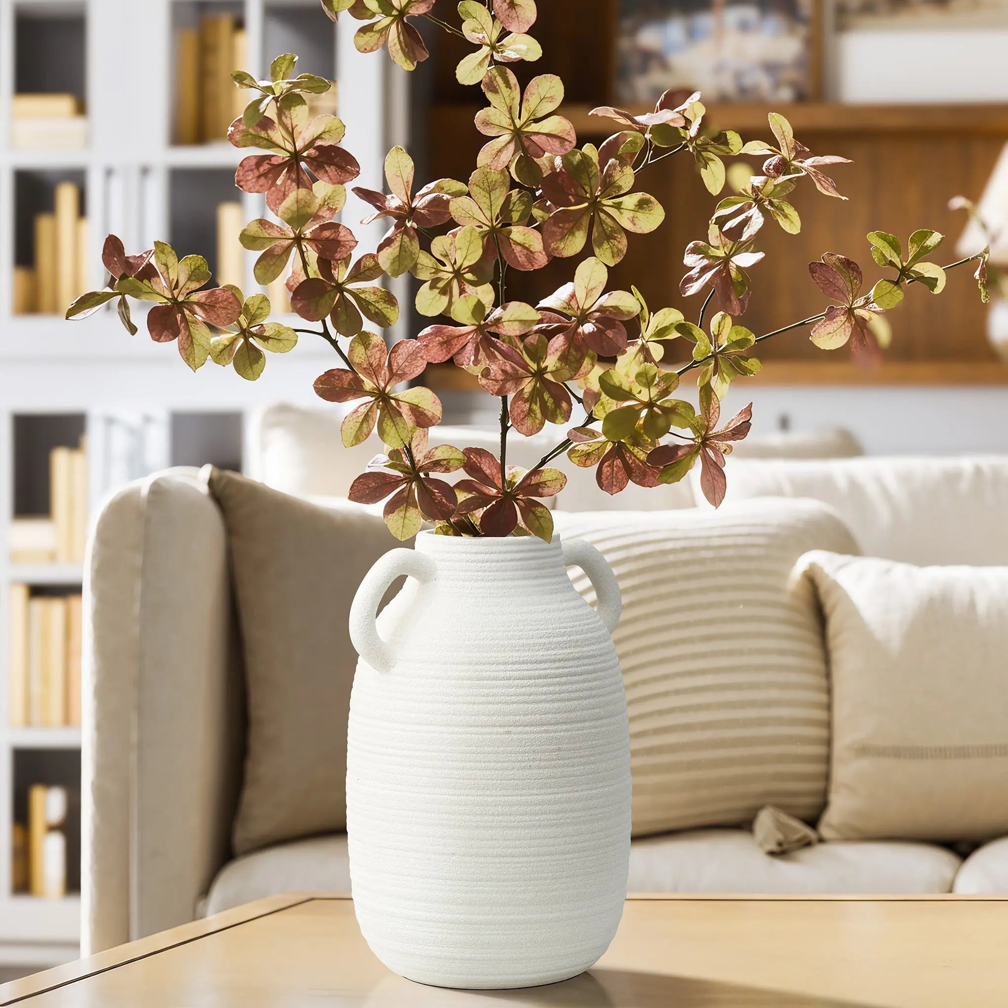 A textured ceramic vase with handles holds a leafy branch with brown and green leaves. It exudes rustic charm as it sits on a wooden table in the cozy living room setting.