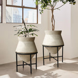 Two large, beige cachepot planters on black metal stands hold tall, leafy plants, adding a touch of vintage distressed style to the sunlit room. They are beautifully placed in front of large windows, enhancing the indoor space decor with their textured charm.